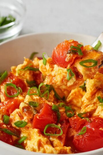 A bowl of Chinese Tomato Egg Stir-Fry over white rice, with soft scrambled eggs, juicy red tomato pieces, and garnished with sliced green onions, with a fork lifting a portion of the dish.