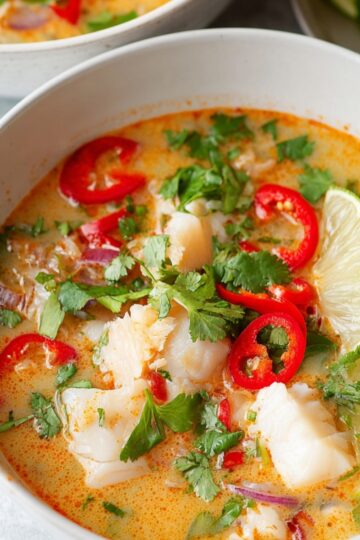 A close-up of a bowl of creamy coconut lime fish soup, featuring white fish pieces, red chili slices, fresh cilantro, and lime wedges. The soup has an orange tint and looks rich, fragrant, and ready to serve.