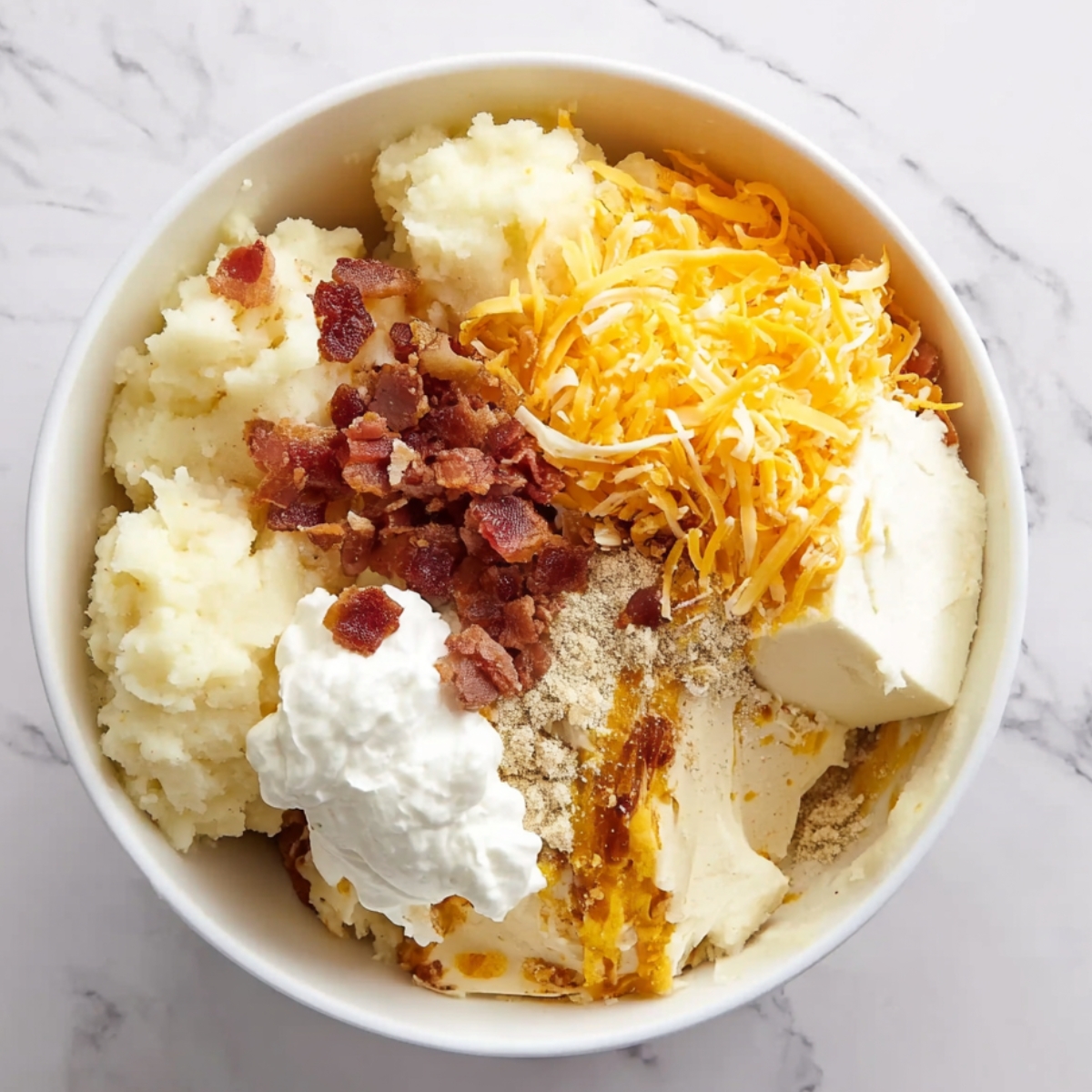 op-down view of a white mixing bowl filled with scooped mashed potatoes, shredded cheddar cheese, crumbled bacon, cream cheese, sour cream, and seasonings, ready to be mixed for a twice-baked potato casserole.