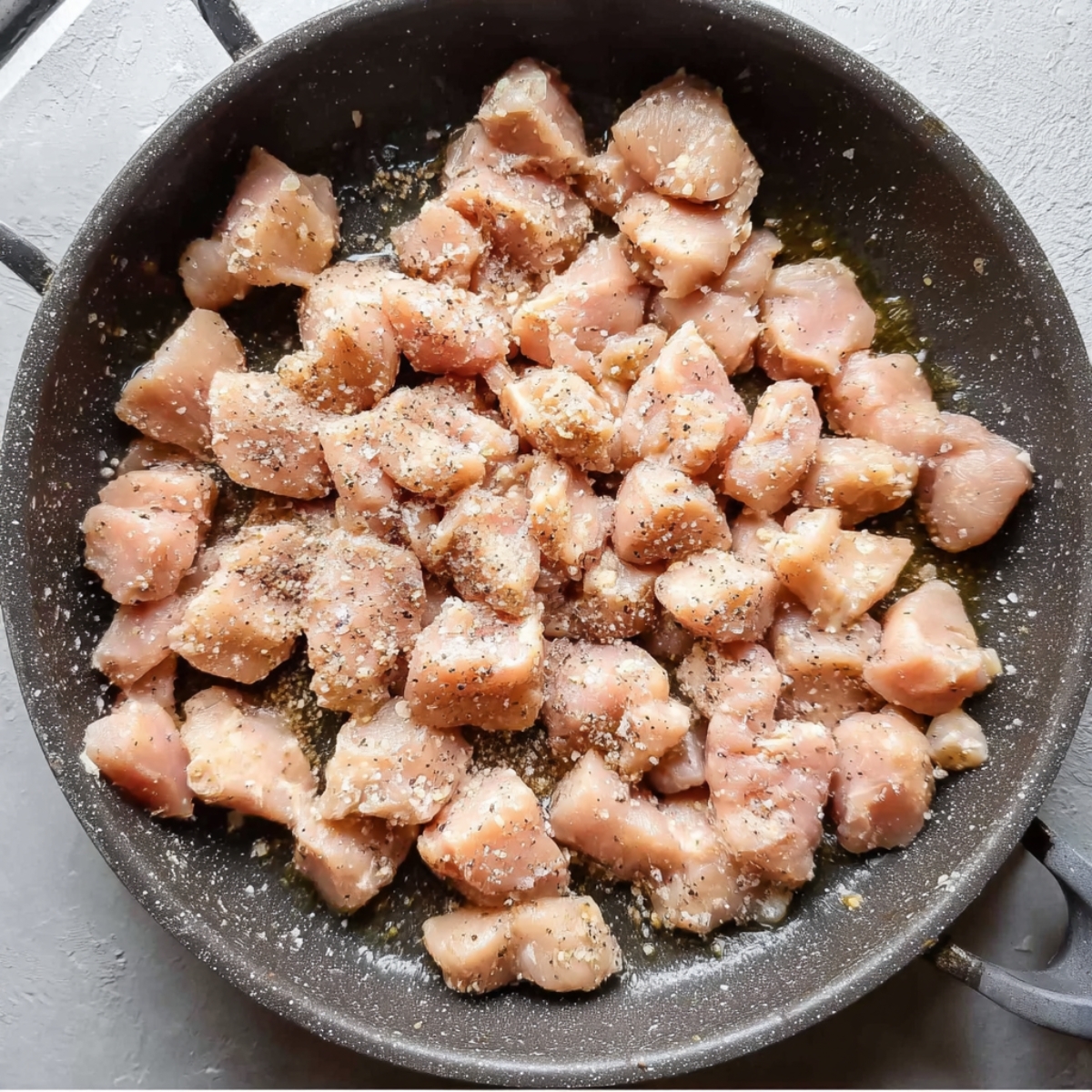 Raw diced chicken pieces in a skillet, lightly seasoned with salt and pepper, ready to be cooked, overhead view, high detail food prep photography.