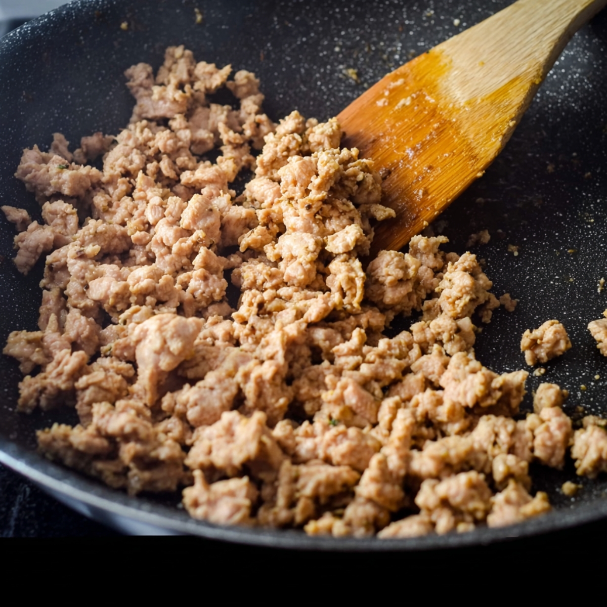 Close-up of seasoned ground chicken cooking in a nonstick skillet, stirred with a wooden spatula, highlighting the texture and browning of the meat.