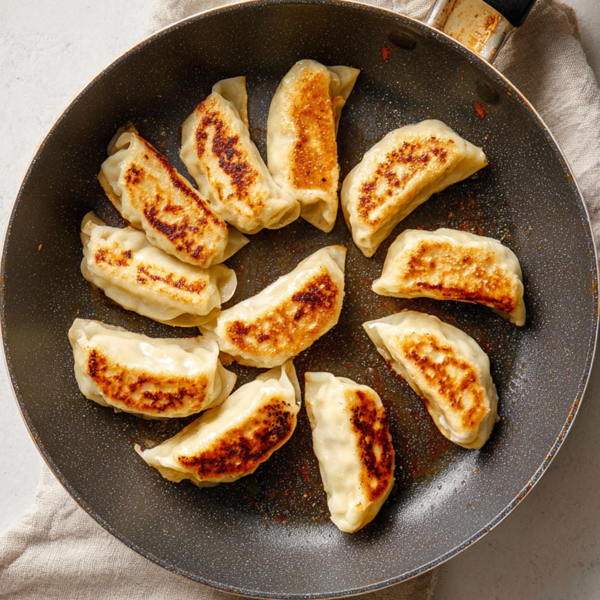Top-down view of a nonstick skillet with golden-brown pan-fried dumplings arranged in a circular pattern, showing crispy bottoms and soft tops, on a light countertop with a neutral cloth beside it.