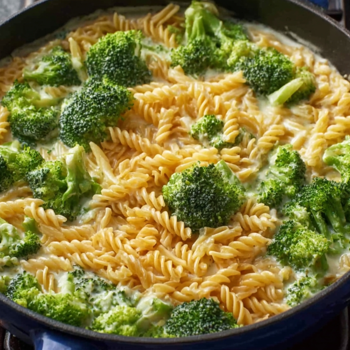 Fusilli pasta and fresh broccoli florets cooking in a creamy sauce in a black skillet, broccoli partially submerged, top-down view, showing vibrant green broccoli and uncooked pasta spirals in a rich white sauce.
