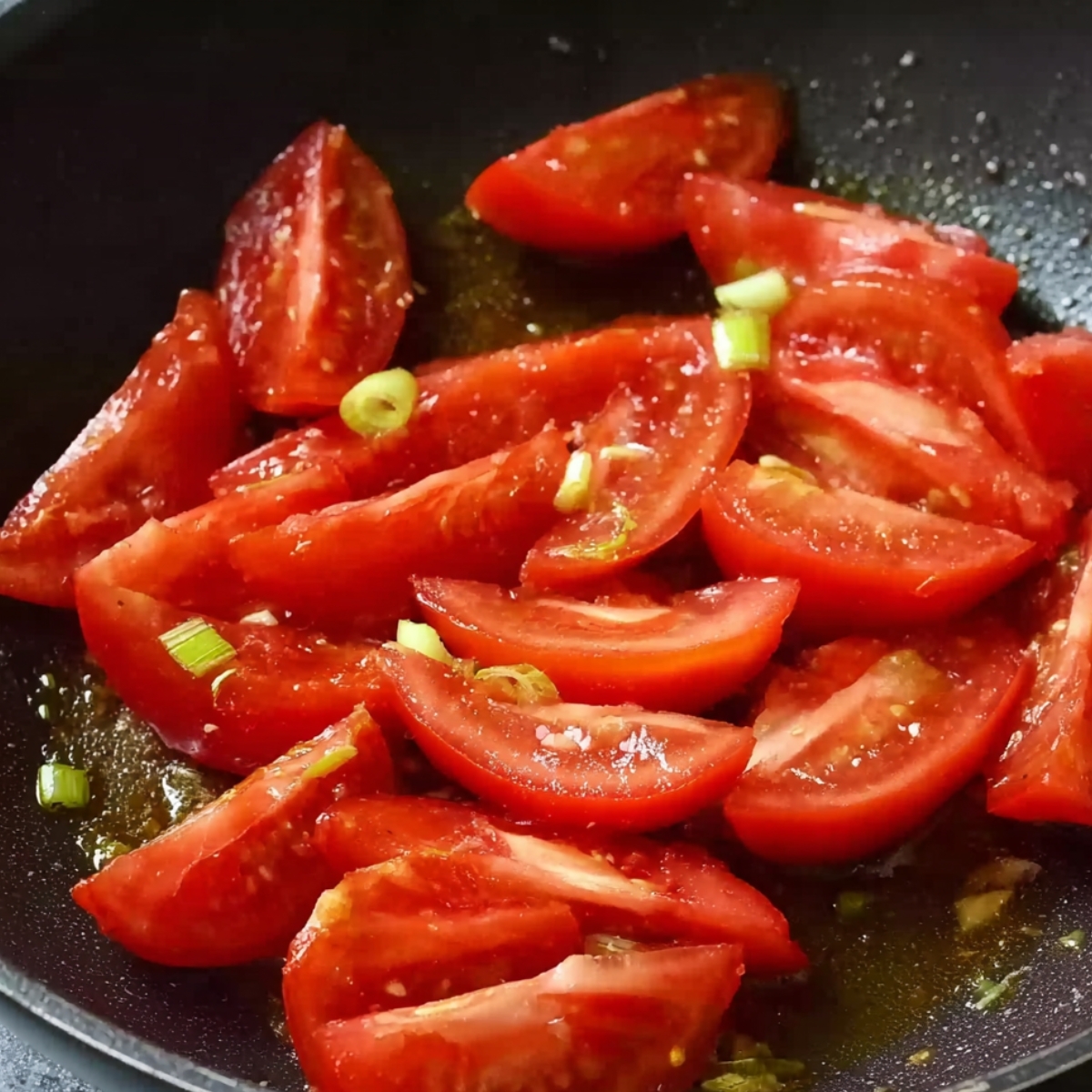 Fresh tomato wedges cooking in a black non-stick pan with a few pieces of chopped green onions, glistening with oil, showing the start of a home-cooked Chinese tomato and egg stir-fry.
