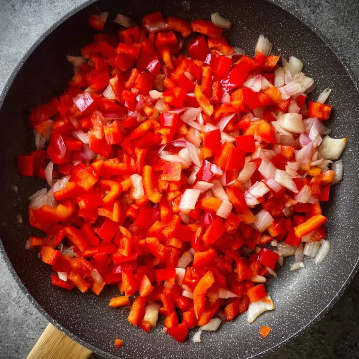 Diced red bell peppers and onions cooking in a skillet, beginning to soften, overhead view, bright natural lighting, close-up of colorful vegetables.
