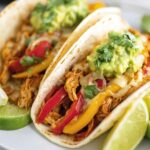 Close-up of Crockpot Chicken Fajitas filled with cooked shredded chicken, sautéed red and yellow bell peppers, onions, melted cheese, and a dollop of guacamole, garnished with chopped cilantro and lime wedges on the side, served on a white plate.