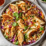 Top-down view of a large round bowl filled with an Dumpling Salad, topped with pan-fried dumplings, shredded cabbage, carrots, purple cabbage, green onions, cilantro, and sesame seeds, with small bowls of dipping sauce visible in the background, on a light-colored countertop.