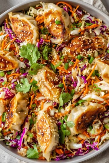 Top-down view of a large round bowl filled with an Dumpling Salad, topped with pan-fried dumplings, shredded cabbage, carrots, purple cabbage, green onions, cilantro, and sesame seeds, with small bowls of dipping sauce visible in the background, on a light-colored countertop.