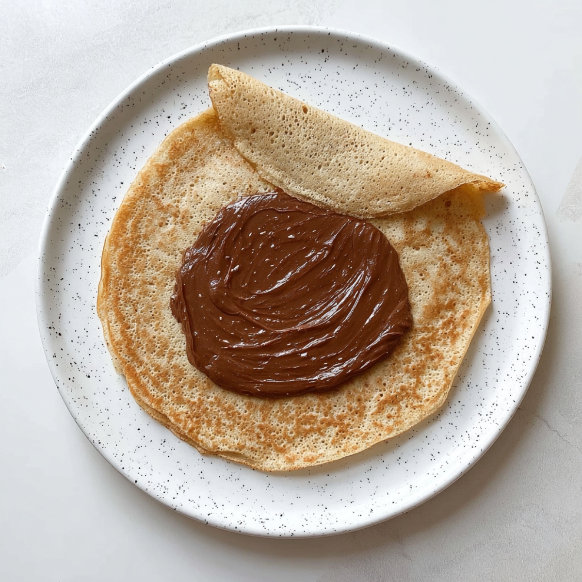 Thin crepe on a white speckled plate with a large spread of chocolate hazelnut filling in the center and one side folded over.