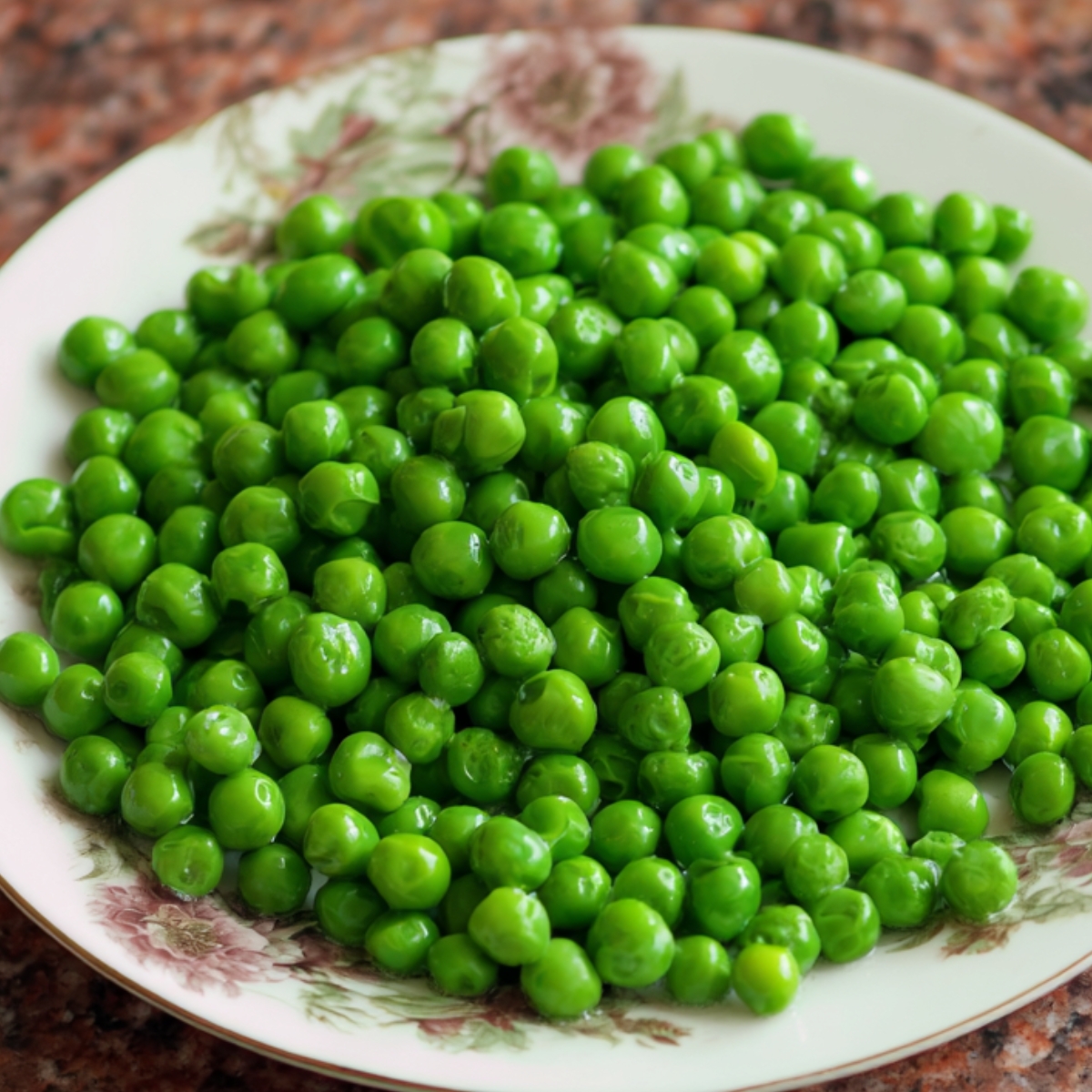 A white plate filled with fresh, bright green peas, arranged neatly. The plate has a subtle floral pattern and sits on a brown granite countertop.
