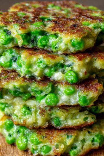 A stack of golden-brown green pea fritters on a wooden cutting board. The fritters are thick, crispy on the outside, and filled with vibrant green peas in a creamy batter.