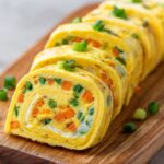 Sliced Japanese rolled omelet (Gyeran Mari) on a wooden board, showing bright yellow layers filled with colorful diced vegetables like carrots and green onions, garnished with extra green onion pieces.