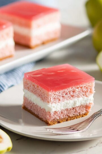 Close-up of a plated slice of Hawaiian Guava Cake with a pink gelatin top layer and a white cream cheese middle layer, served on a white plate with a silver fork, with more cake slices on a rectangular plate and fresh guavas in the background.