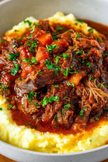Another close-up of Italian Pot Roast on mashed potatoes, with tender meat in tomato-based sauce, diced carrots, and fresh parsley on top, served in a white bowl.
