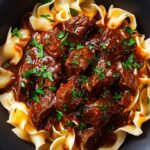 A close-up of a bowl of Korean Beef Noodles. Tender beef chunks in a dark, savory sauce sit atop egg noodles, garnished with fresh chopped herbs, creating a rich and appetizing presentation.