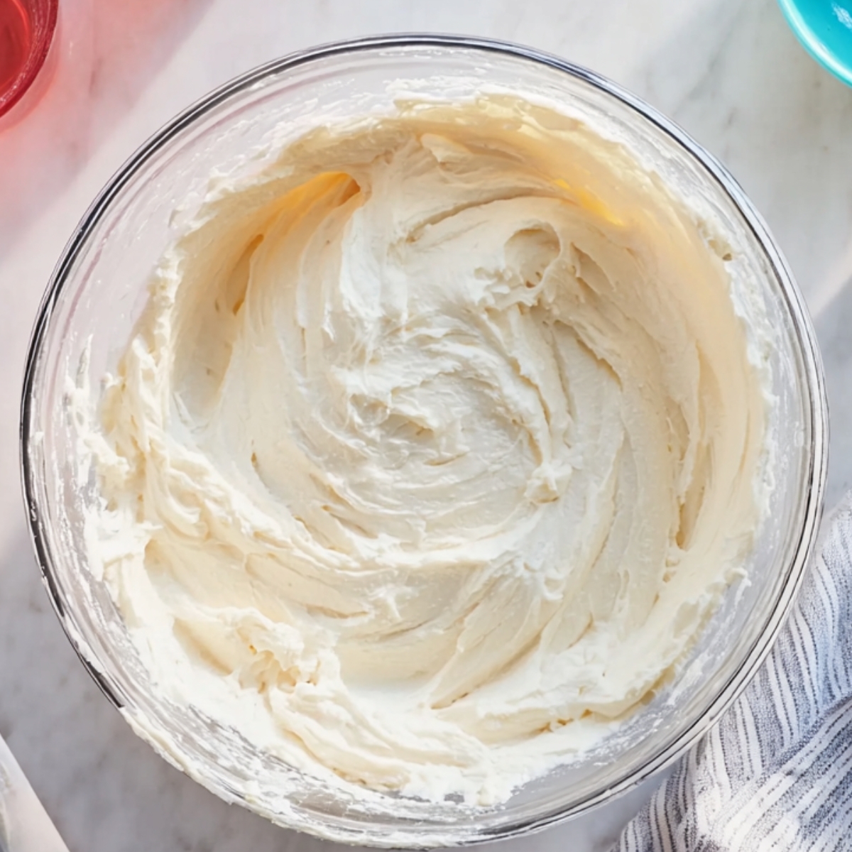 Top-down view of a clear glass bowl filled with smooth, whipped cream cheese frosting, placed on a marble countertop with a striped cloth and a small turquoise bowl partially visible.