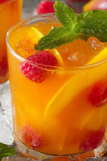A glass of mango iced tea with ice cubes, fresh raspberries, mango slices, and a sprig of mint on top, surrounded by additional ice and mint leaves.
