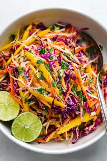 Overhead view of a white bowl with colorful mango slaw, including julienned mango, shredded purple cabbage, carrots, and chopped herbs, garnished with two lime halves and a silver serving spoon.