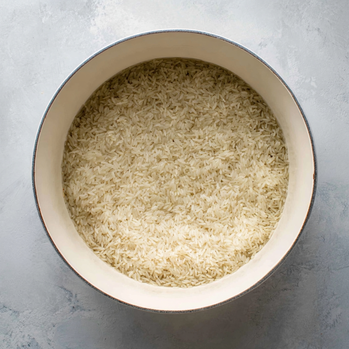 Top-down view of a white pot filled with uncooked long-grain rice, evenly spread. Neutral background, showing the individual rice grains clearly.