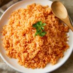 A white plate with cooked Authentic Mexican Rice, fluffy and reddish-orange in color, garnished with a small sprig of fresh parsley on top. A wooden spoon rests on the side of the plate.