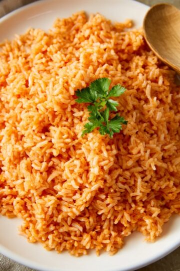 A white plate with cooked Authentic Mexican Rice, fluffy and reddish-orange in color, garnished with a small sprig of fresh parsley on top. A wooden spoon rests on the side of the plate.