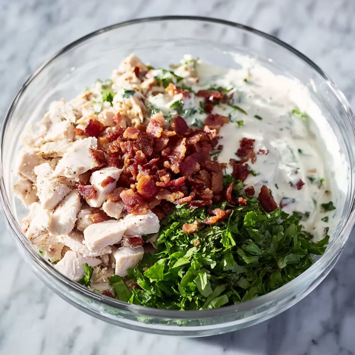 Glass bowl with chopped cooked chicken, crispy bacon bits, fresh chopped greens, and creamy ranch dressing, ingredients unmixed, on a marble countertop, bright natural lighting.