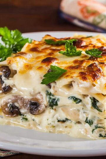 Baked creamy mushroom and spinach lasagna in a white ceramic baking dish, golden melted cheese on top with browned spots, sprinkled with chopped parsley, on a wooden table with an orange and white checkered kitchen towel beside it.