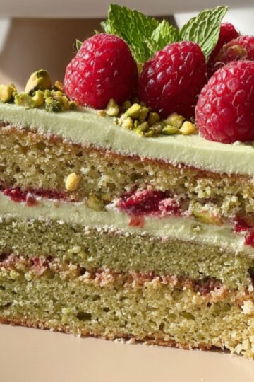 A close-up slice of Pistachio Raspberry Cake with green frosting, layers of raspberry jam, and chopped pistachios. The top is garnished with fresh raspberries and mint leaves.