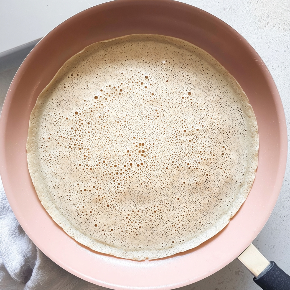 Fresh crepe cooking in a non-stick frying pan, showing small bubbles across the surface as it cooks.