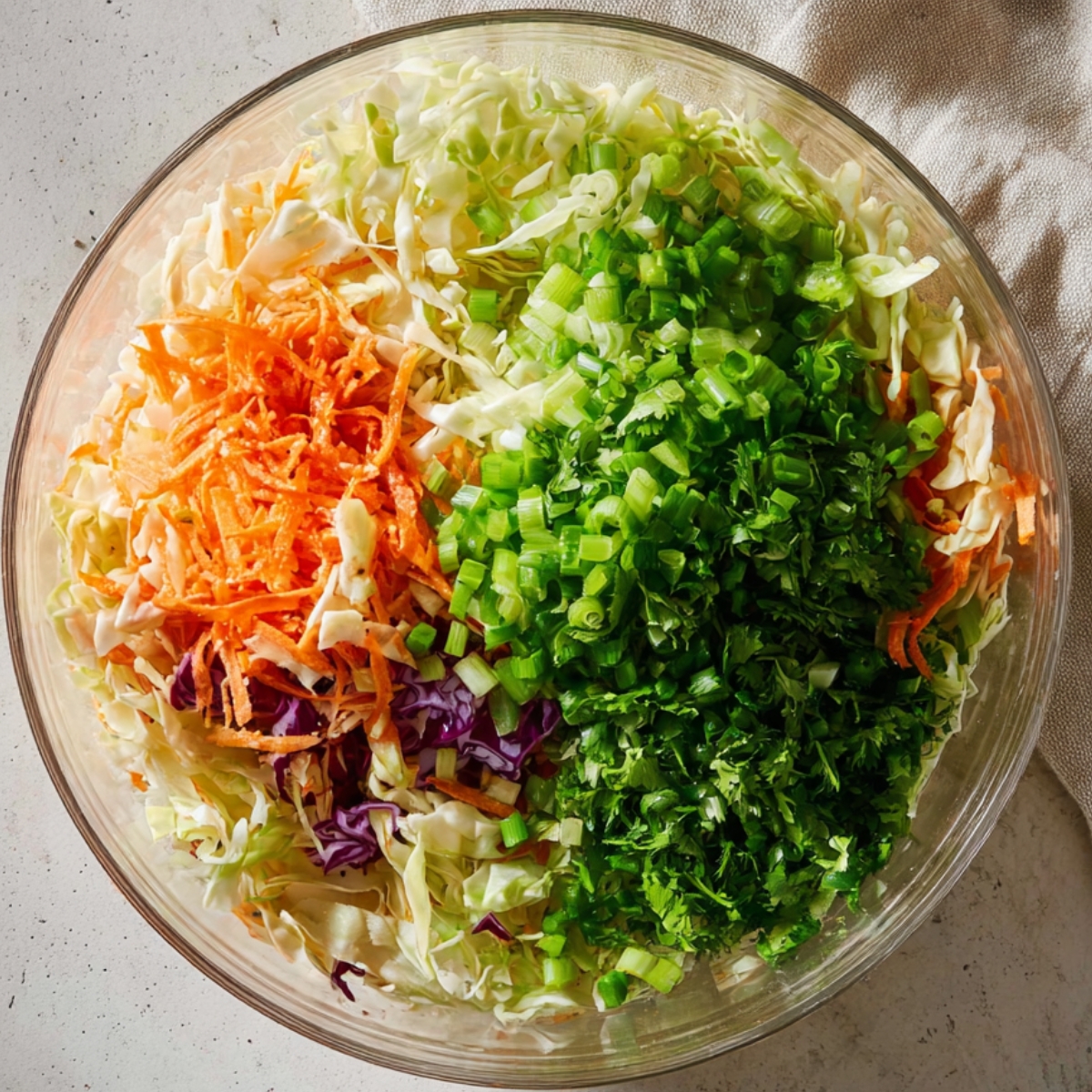Top-down view of a clear glass bowl filled with freshly chopped salad ingredients, including shredded cabbage, grated carrots, chopped green onions, and fresh herbs, on a light countertop with a neutral cloth nearby.