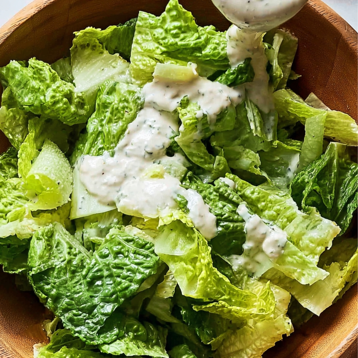 Fresh green romaine lettuce salad in a wooden bowl being topped with creamy dressing from a spoon.
