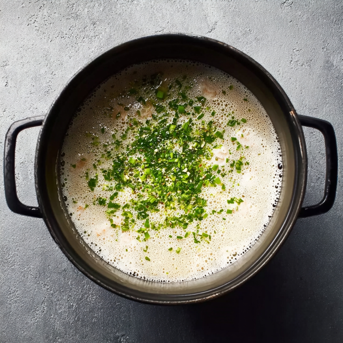 Close-up overhead view of a black pot with the base of seafood bisque, creamy light mixture with small bits of vegetables and herbs, foamy texture, natural light, cooking preparation stage.