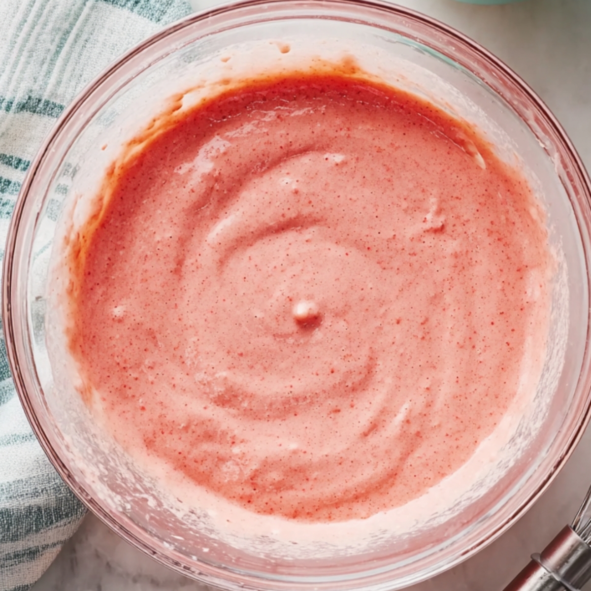 Top-down view of a glass bowl containing pink guava cake batter, with a whisk resting on the side and a striped kitchen towel in the background, bright natural lighting highlighting the smooth texture.