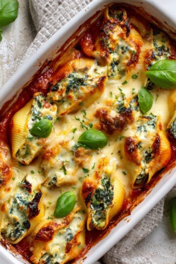 Baked Ricotta and Spinach Stuffed Shells in a white rectangular dish, topped with melted cheese and garnished with fresh basil leaves, with a rustic textured background and a light-colored napkin.