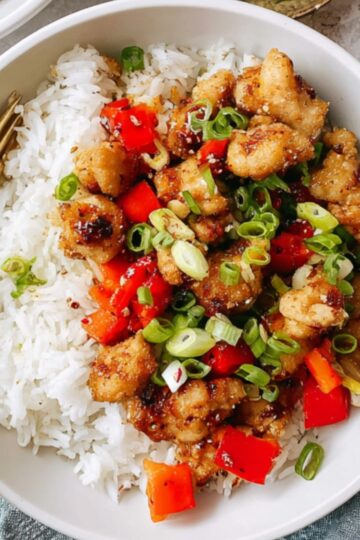 A bowl of cooked Salt And Pepper Chicken served over white rice, topped with diced red bell peppers and sliced green onions, gold fork in the bowl, overhead view, bright natural lighting, appetizing Asian-style meal.
