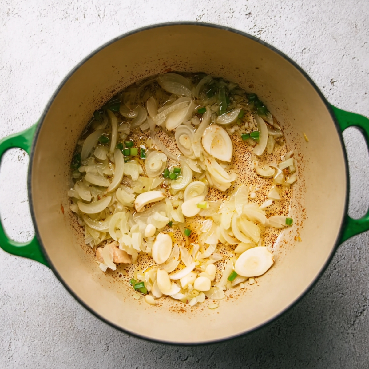 A green-handled pot with sliced onions, garlic, and chopped green chilies sautéing in oil. The ingredients are lightly browned, releasing aromatic flavors, and the pot shows a hint of seasoning residue on the bottom.