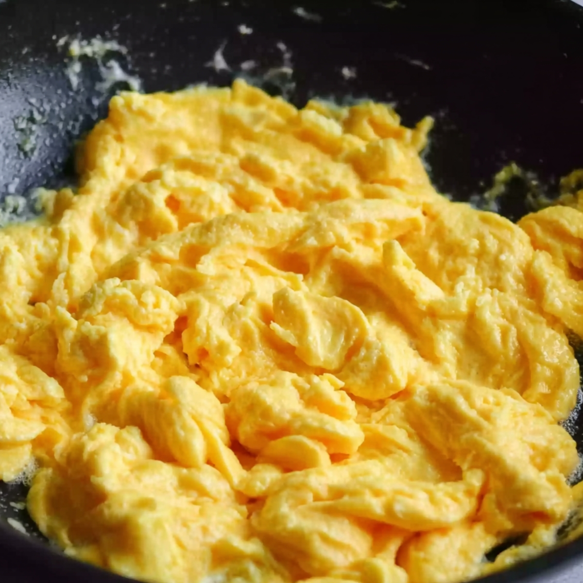 Fluffy, soft scrambled eggs cooking in a black non-stick pan, with a creamy yellow texture, forming curds as they cook.