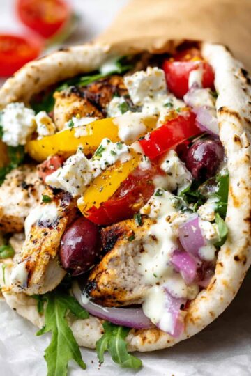 Close-up of a Mediterranean-style Sheet Pan Chicken Pitas filled with grilled chicken, red and yellow bell peppers, cherry tomatoes, red onions, olives, arugula, and crumbled feta cheese, drizzled with creamy sauce, on white parchment with cherry tomatoes and arugula leaves in the background.
