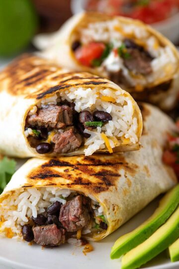 Close-up of a sliced Steak Burrito on a plate, filled with steak, rice, black beans, and melted cheese, with a side of pico de gallo and sliced avocado.