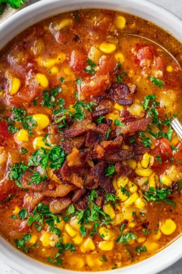 White bowl of tomato corn chowder garnished with crispy bacon, chopped parsley, and corn, with a spoon partially dipped in the soup, bright and appetizing presentation.