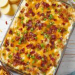 Top-down view of a glass baking dish filled with Twice Baked Potato Casserole, topped with melted cheddar cheese, crispy bacon bits, and sliced green onions, ready to serve on a white wooden surface.