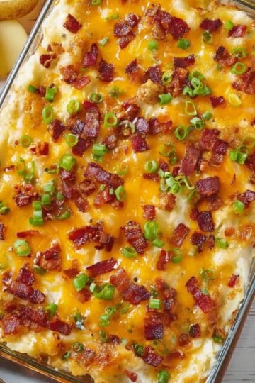 Top-down view of a glass baking dish filled with Twice Baked Potato Casserole, topped with melted cheddar cheese, crispy bacon bits, and sliced green onions, ready to serve on a white wooden surface.