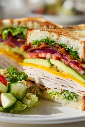 A stacked Ultimate Club Sandwich on a plate with toasted bread, layers of turkey, cheddar cheese, avocado, tomato, red onion, bacon, and lettuce. Served alongside a fresh salad of cherry tomatoes, cucumber, and leafy greens.