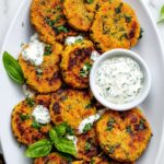 Cooked golden-brown Vegetable Chickpea Fritters arranged on a white plate, garnished with fresh basil and parsley, served with a small bowl of creamy herb yogurt sauce, rustic flat-lay view on a white surface, appetizing vegan dish, high-resolution, warm lighting.