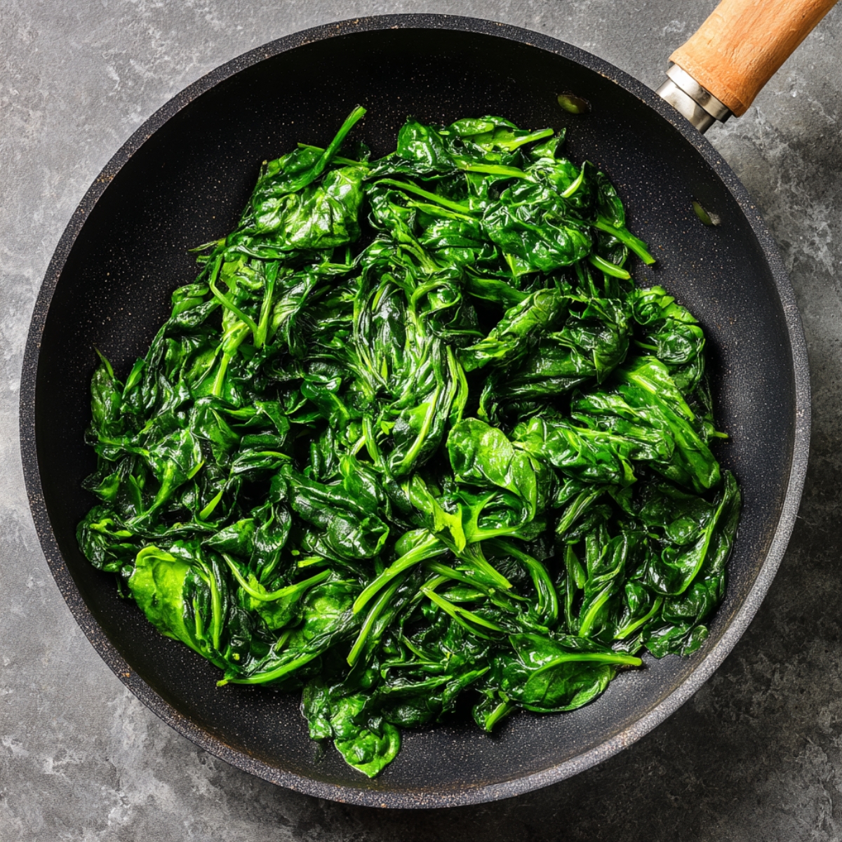 Fresh spinach leaves lightly sautéed in a black frying pan, showing a glossy, wilted texture and vibrant green color.
