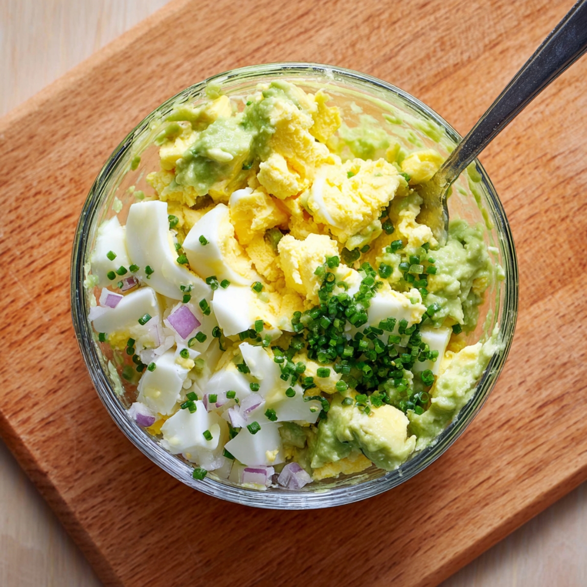 A glass bowl filled with chopped hard-boiled eggs, mashed avocado, red onion, and chives, partially mixed on a wooden board.