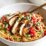 A white bowl of cooked bruschetta chicken pasta topped with sliced grilled chicken, diced tomatoes, and fresh basil, showcasing a colorful and appetizing Bruschetta Chicken Pasta meal.