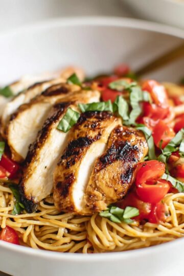 A white bowl of cooked bruschetta chicken pasta topped with sliced grilled chicken, diced tomatoes, and fresh basil, showcasing a colorful and appetizing Bruschetta Chicken Pasta meal.