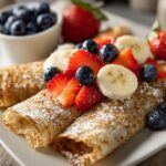 Rolled oatmeal crepes topped with sliced strawberries, banana, blueberries, and powdered sugar on a white plate.